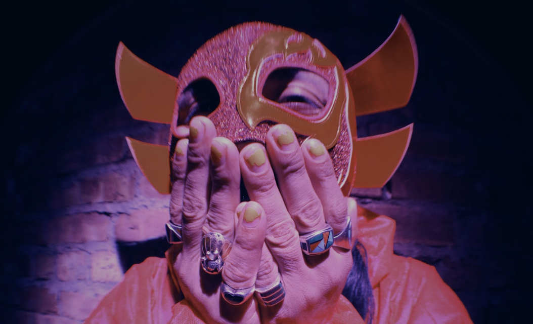 Imagen: Un hombre con la mascara roja de Lucha Lupe puesta con las manos sobre la boca con uñas pintadas color amarillo.